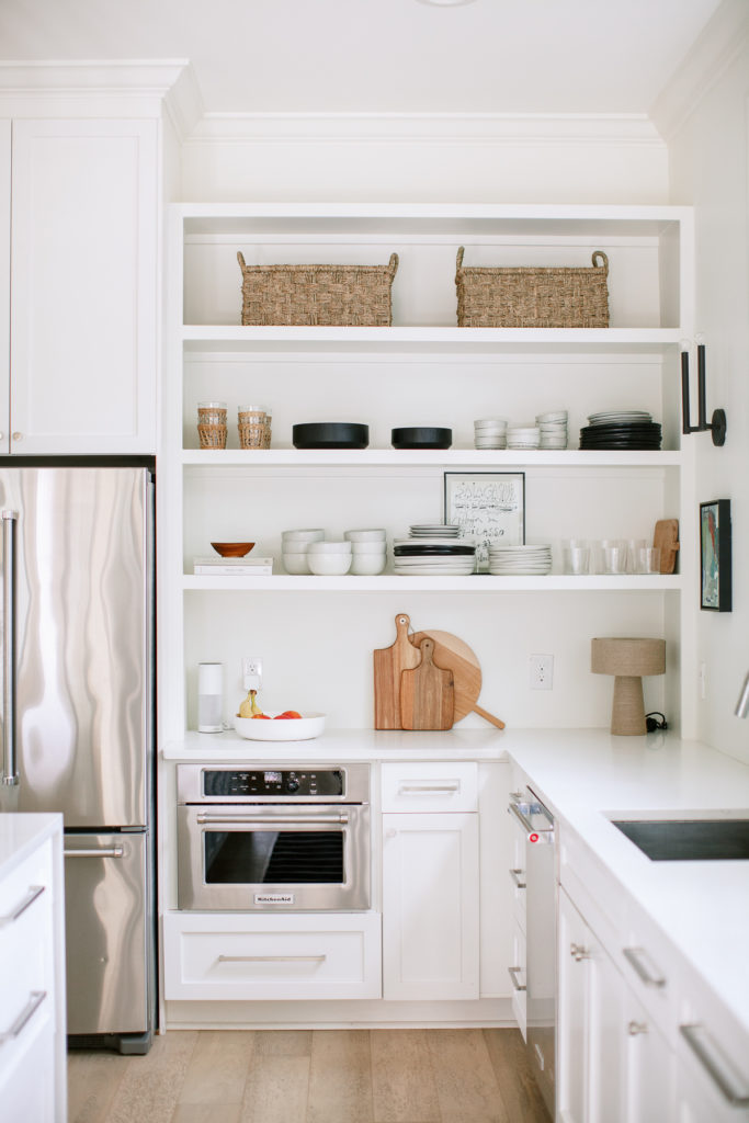 white kitchen with open shelving that has dishes and glasses neatly organized, baskets above, and wood cutting boards stacked underneath