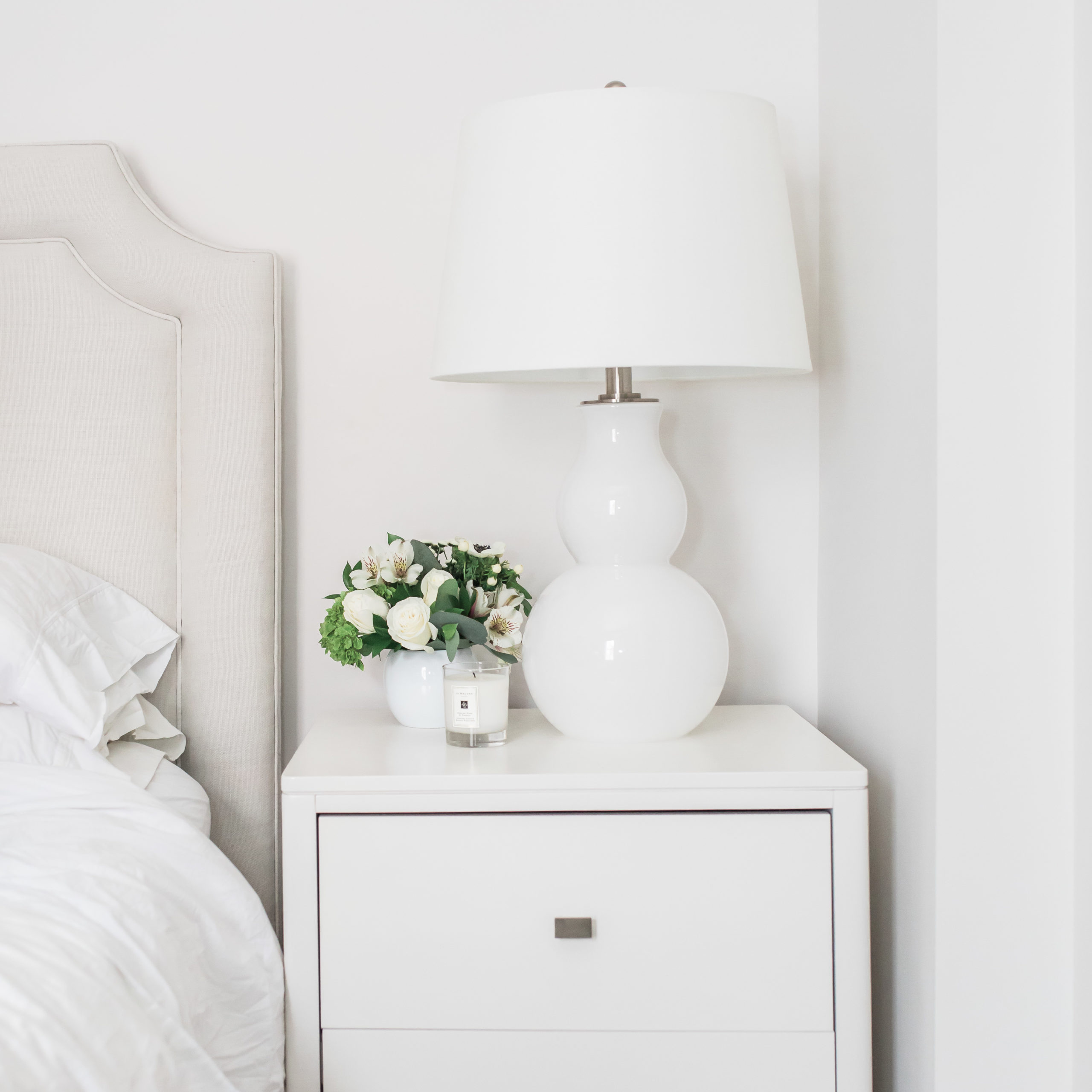 corner of a bedroom with a white nightstand, lamp, and pretty flower arrangement