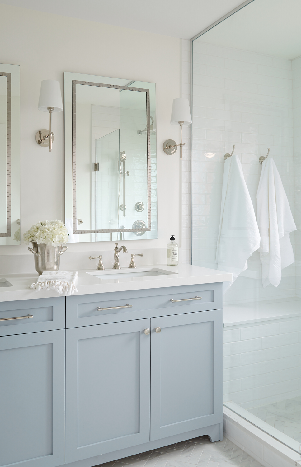 corner of bathroom showing light blue vanity with polished nickel faucets and hardware next to walk in shower with white fluffy towels hanging inside