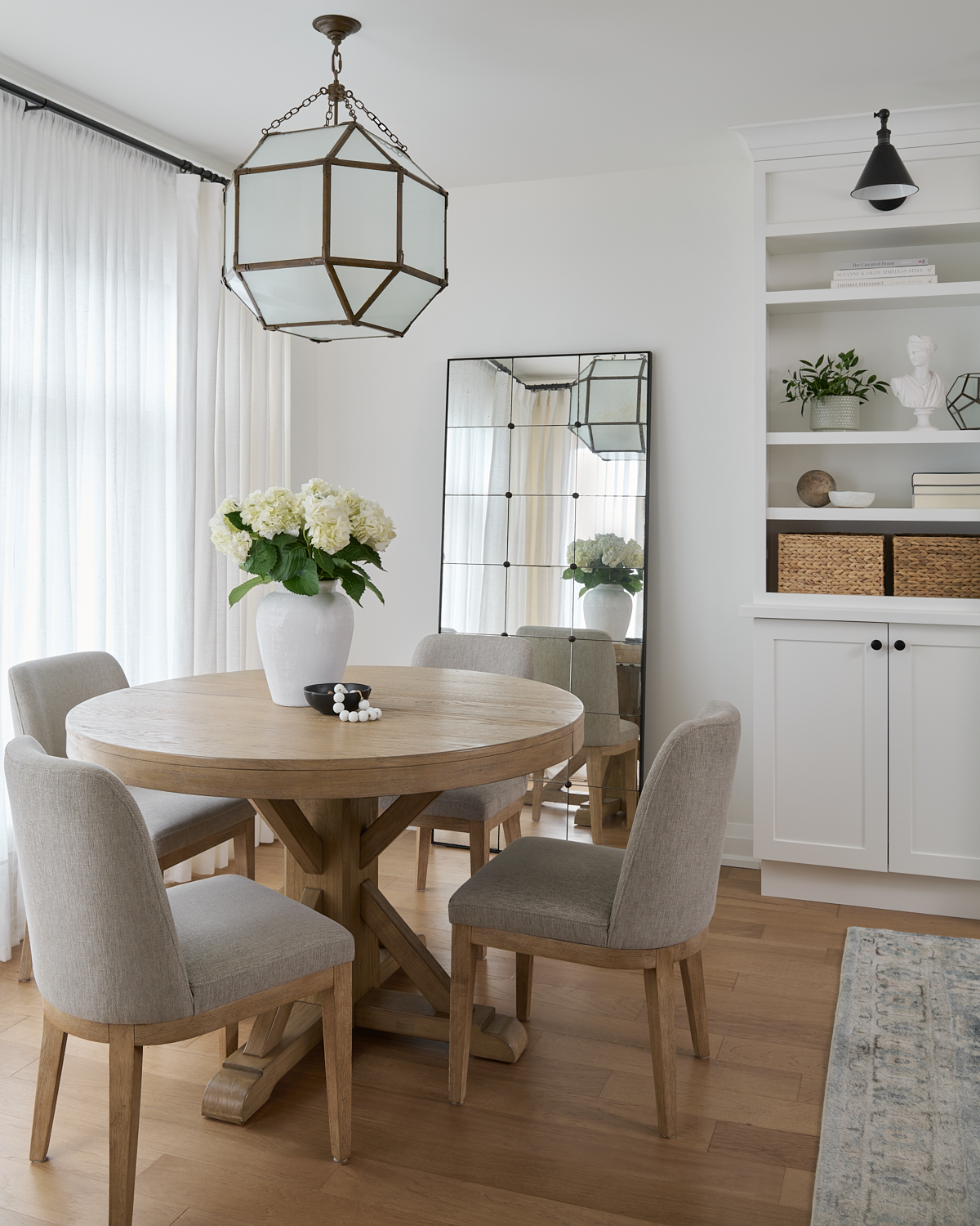 Dining room image with white oak round dining table surrounded with neutral upholstered chairs. White linen drapery behind and a wrought iron light fixture above.