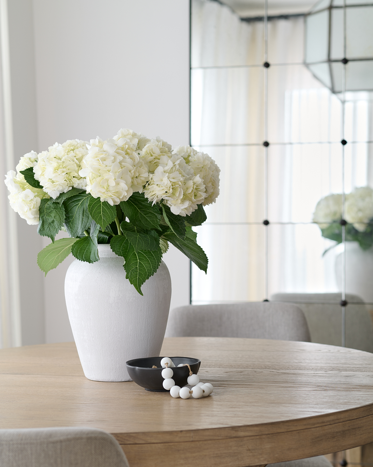 White hydrangeas in white vase on wooden dining table with dark bowl and white beads next to it
