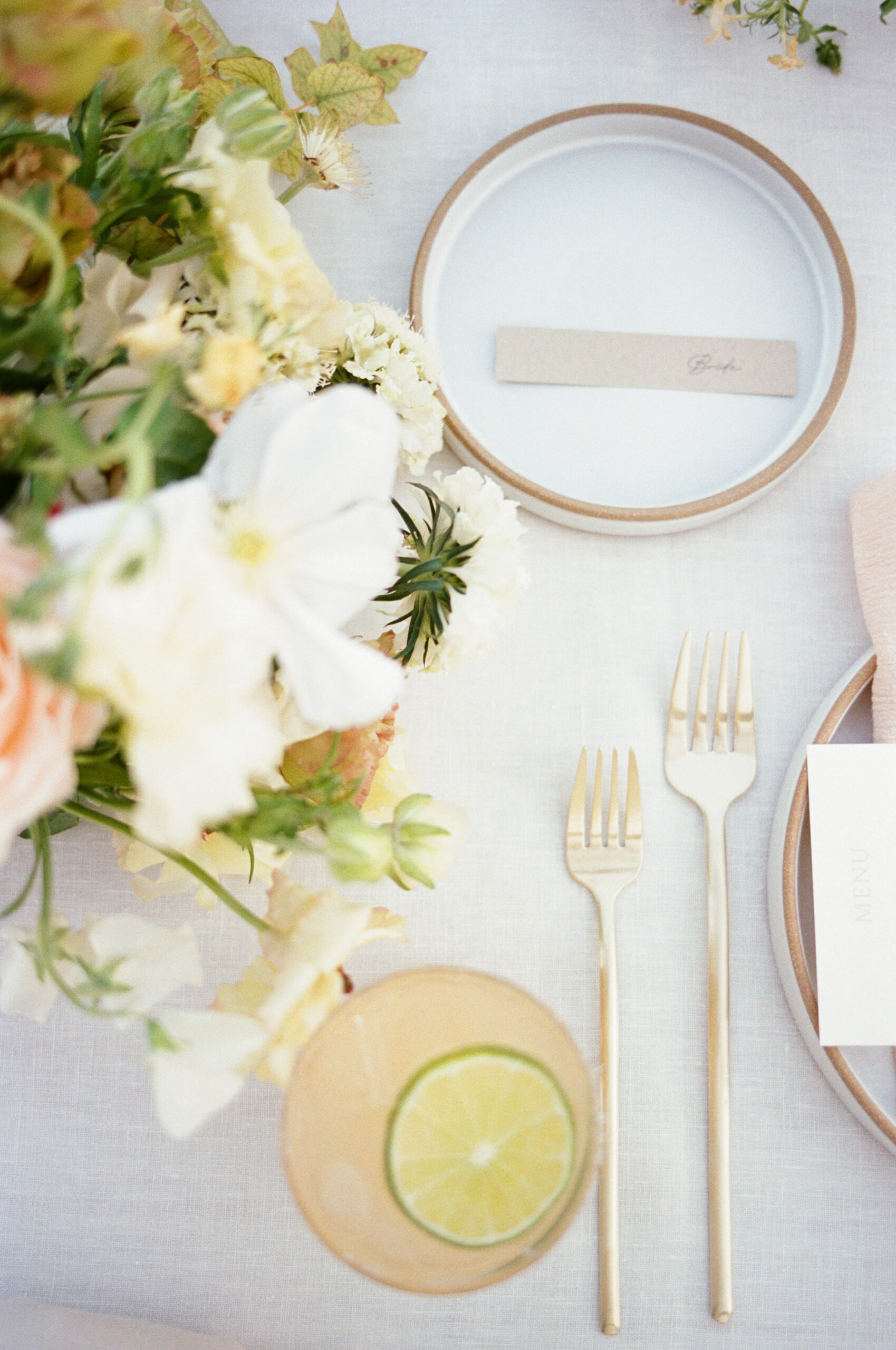 table top with floral arrangement, peach coloured cocktail with lime floating inside, and white plates with gold cutlery next to them