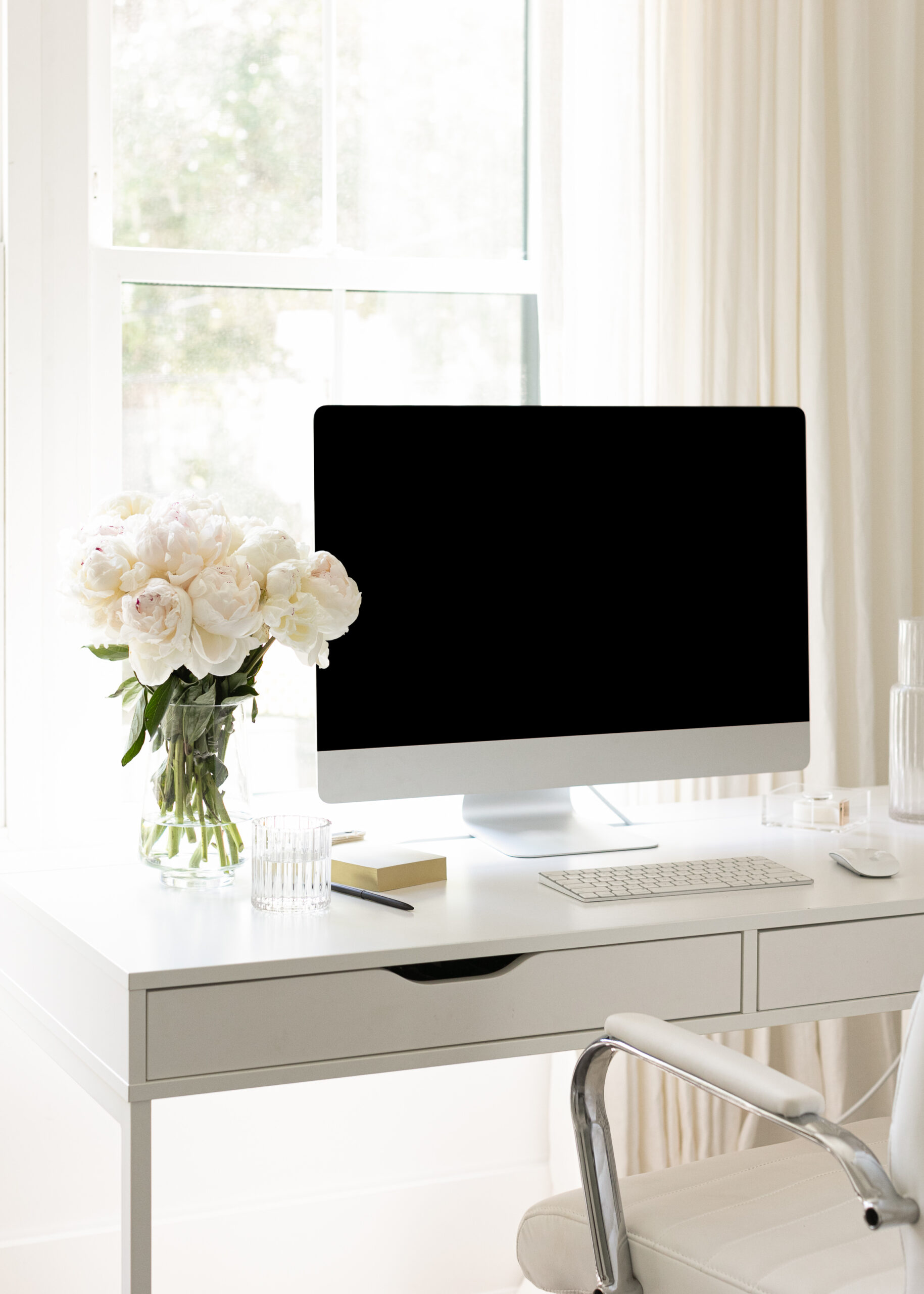 White desk in front of a window with a vase of peonies next to a computer