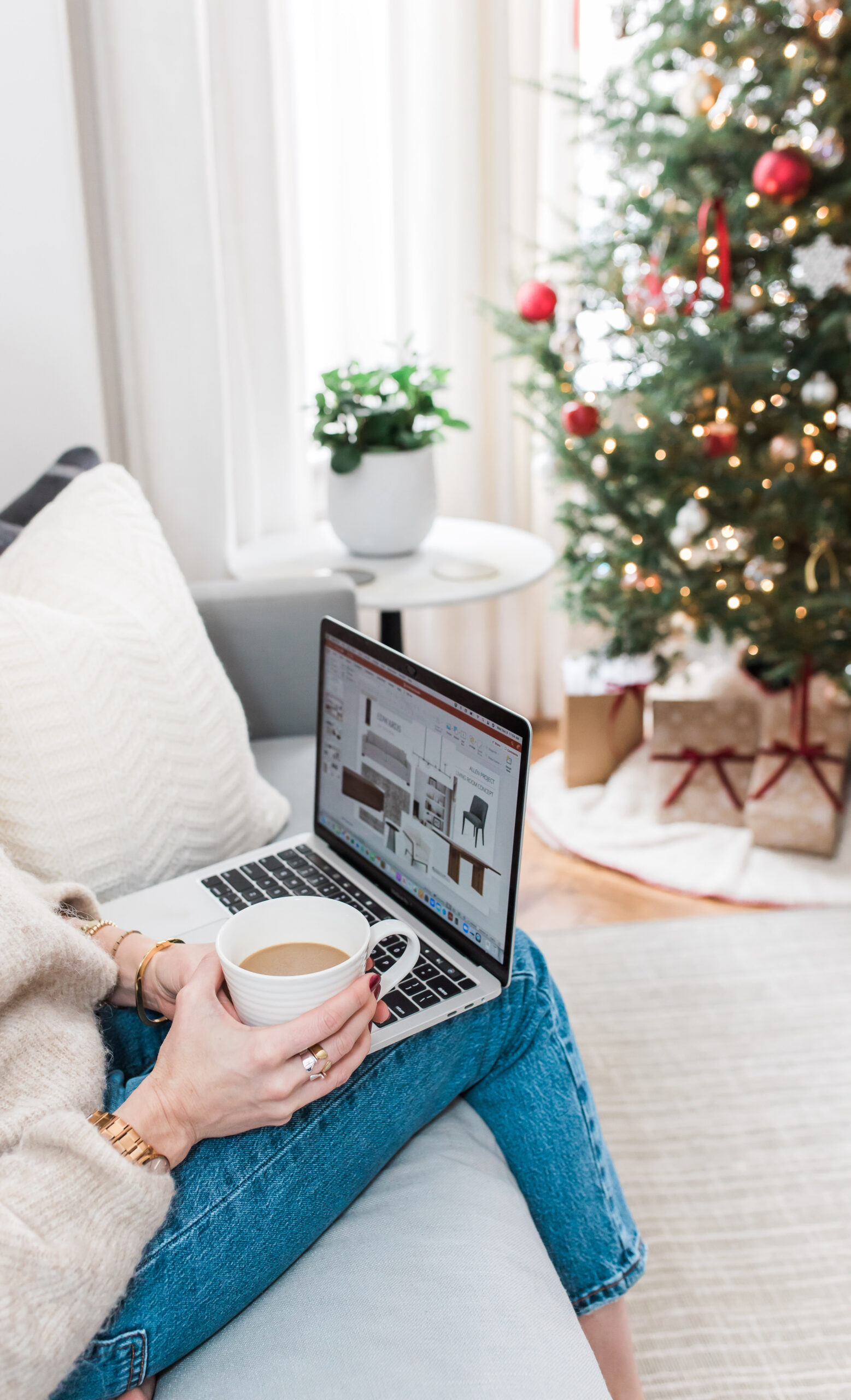 lady with laptop on her lap and coffee in hand with Christmas tree and gifts in the background
